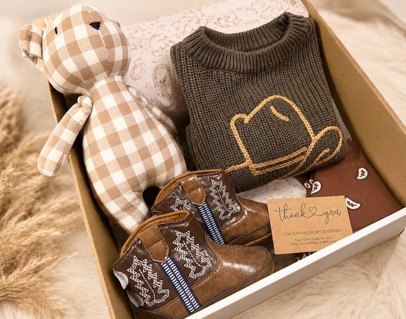May include: A gift box containing a beige and white gingham teddy bear, a dark olive green knit sweater with a yellow embroidered cowboy hat outline, brown faux leather cowboy boots with white stitching, and a thank you card with text.
