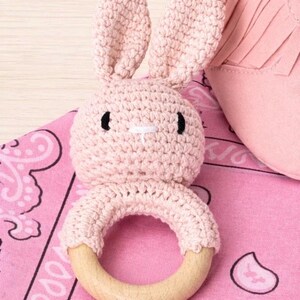 May include: A handmade pink crochet bunny rattle with a wooden ring base. The bunny has black eyes and a white cross-stitch nose. The rattle is on a pink paisley patterned fabric, with a pink fringed item in the background.