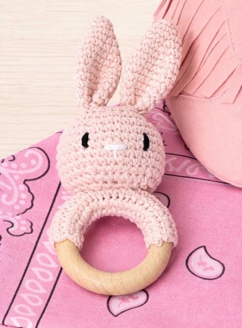 May include: A handmade pink crochet bunny rattle with a wooden ring base. The bunny has black eyes and a white cross-stitch nose. The rattle is on a pink paisley patterned fabric, with a pink fringed item in the background.