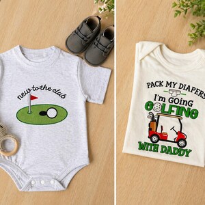 May include: Two baby onesies with golf-themed designs. One is light gray with "new to the club" text and a golf course graphic. The other is cream-colored with "Pack my diapers I'm going golfing with Daddy" text and a golf cart graphic. Gray baby shoes and a toy are also present.