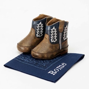 Pode incluir: Um par de botas de cowboy castanhas e azul marinho para beb&eacute; com costuras brancas. As botas est&atilde;o sobre uma bandana azul marinho com o nome "Rome" bordado em branco.