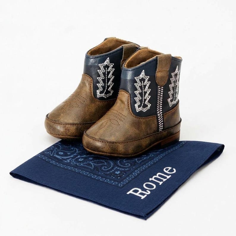 Pode incluir: Um par de botas de cowboy castanhas e azul marinho para beb&eacute; com costuras brancas. As botas est&atilde;o sobre uma bandana azul marinho com o nome "Rome" bordado em branco.