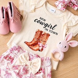 A cream-coloured pink ruffled baby outfit set with a "New Cowgirl in Town" graphic, featuring cowboy boots. The set includes a onesie, shorts, two headbands, pink fringed booties, and a pink bunny rattle. The shorts have a bow. baby girl gift box ideas. pink cowgirl gifts set. Texas baby girl gifts. southern baby girl gift. farm baby girl gift. affordable baby girl gift. Oklahoma baby girl gift. Montana baby girl gift. Wyoming baby girl gift. Indian girl gift. baby shower gift. baby blanket