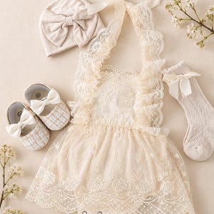 May include: A cream-colored baby outfit set featuring a lace romper, a matching hat with a bow, socks with a bow, and a pair of shoes with bows. The outfit is designed with delicate lace details and a soft, neutral color palette.