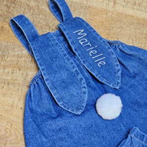 A blue denim overall with bunny ear detailing. The ears are embroidered with the name "Marielle" in white. A fluffy white pom-pom is attached to the back, resembling a bunny tail. The overall is designed for a child. Cute bunny Easter overall