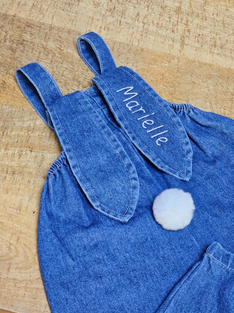A blue denim overall with bunny ear detailing. The ears are embroidered with the name "Marielle" in white. A fluffy white pom-pom is attached to the back, resembling a bunny tail. The overall is designed for a child. Cute bunny Easter overall
