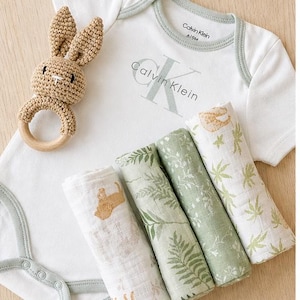 May include: A baby gift set featuring a white onesie with the Calvin Klein logo, a crochet bunny rattle, and four rolled muslin swaddles with nature-themed prints. The onesie is size 6/9M.