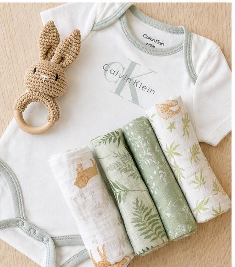 May include: A baby gift set featuring a white onesie with the Calvin Klein logo, a crochet bunny rattle, and four rolled muslin swaddles with nature-themed prints. The onesie is size 6/9M.