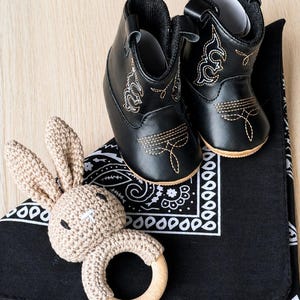 May include: A pair of black baby cowboy boots with gold stitching, a tan crochet bunny rattle, and a black bandana with a white paisley pattern. The boots are made of leather and have decorative stitching.