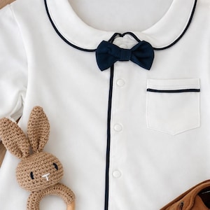 baby gentleman gift set bow onesie. White baby romper with navy blue trim and bow tie. Includes a small pocket and button-up front. A brown crochet bunny teether and brown leather baby shoes are also pictured. little gentleman baby suit gift. baby formal wear. peeky blinders baby gift. baby boy gift ideas. baby bow tie