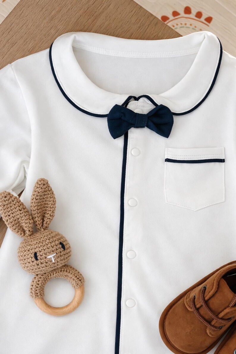 baby gentleman gift set bow onesie. White baby romper with navy blue trim and bow tie. Includes a small pocket and button-up front. A brown crochet bunny teether and brown leather baby shoes are also pictured. little gentleman baby suit gift. baby formal wear. peeky blinders baby gift. baby boy gift ideas. baby bow tie