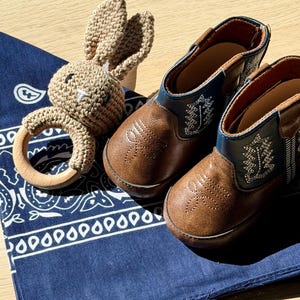 Brown leather baby cowboy boots with decorative stitching and blue detailing, alongside a crocheted bunny rattle and a navy blue paisley bandana. The boots are approximately 10 cm long. Texas themed baby gift. Oklahoma baby gifts. Michigan baby gifts. southern baby gifts. affordable baby gifts. baby gift under 50 dollars. western baby. Mexican baby, country baby keepsake, new baby gift boy. custom baby cowboy gift. custom baby gift. embroidered baby gift. baby cowboy boots. baby boots cowboy.