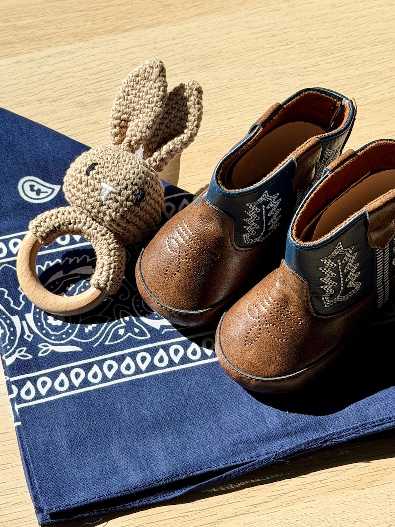 Brown leather baby cowboy boots with decorative stitching and blue detailing, alongside a crocheted bunny rattle and a navy blue paisley bandana. The boots are approximately 10 cm long. Texas themed baby gift. Oklahoma baby gifts. Michigan baby gifts. southern baby gifts. affordable baby gifts. baby gift under 50 dollars. western baby. Mexican baby, country baby keepsake, new baby gift boy. custom baby cowboy gift. custom baby gift. embroidered baby gift. baby cowboy boots. baby boots cowboy.