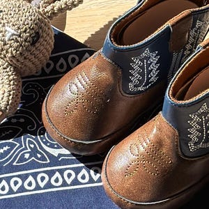 May include: A pair of brown leather baby cowboy boots with decorative stitching and blue accents. A crocheted bunny toy rests beside the boots on a blue bandana with a white paisley pattern. The boots are designed for infants. western baby. Mexican baby, country baby keepsake, new baby gift boy. custom baby cowboy gift. custom baby gift. embroidered baby gift. baby cowboy boots. baby boots cowboy. hunting baby gift. fishing baby gift. fishing enthusiast baby gift. custom baby bandana.