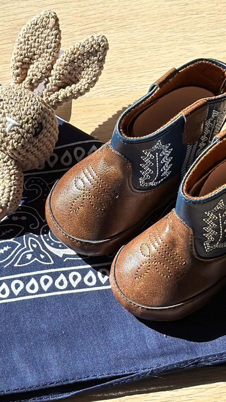May include: A pair of brown leather baby cowboy boots with decorative stitching and blue accents. A crocheted bunny toy rests beside the boots on a blue bandana with a white paisley pattern. The boots are designed for infants. western baby. Mexican baby, country baby keepsake, new baby gift boy. custom baby cowboy gift. custom baby gift. embroidered baby gift. baby cowboy boots. baby boots cowboy. hunting baby gift. fishing baby gift. fishing enthusiast baby gift. custom baby bandana.