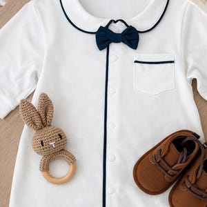baby gentleman gift set bow onesie. White baby romper with navy blue trim and bow tie. Includes a small pocket and button-up front. A brown crochet bunny teether and brown leather baby shoes are also pictured. little gentleman baby suit gift. baby formal wear. peeky blinders baby gift. baby boy gift ideas. baby bow tie
