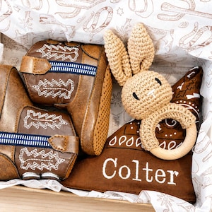 May include: A pair of brown faux leather cowboy boots with white stitching and blue striped accents, a beige crocheted rabbit rattle with a wooden ring, and a brown fabric triangle bib with the white embroidered name Colter in a box.