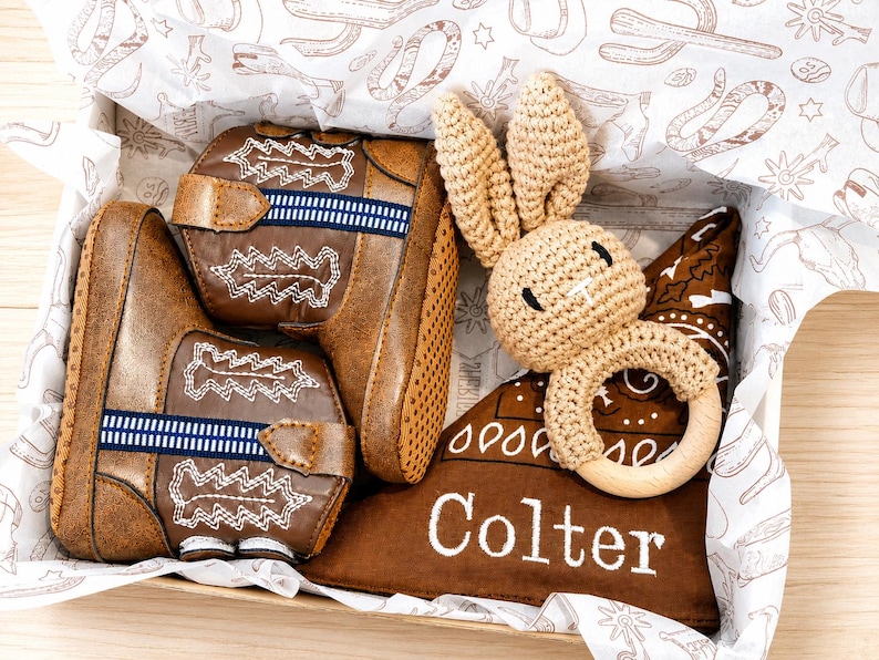 May include: A pair of brown faux leather cowboy boots with white stitching and blue striped accents, a beige crocheted rabbit rattle with a wooden ring, and a brown fabric triangle bib with the white embroidered name Colter in a box.