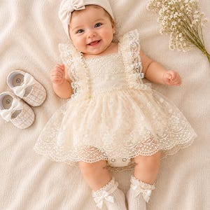 May include: A smiling baby wearing a cream-colored lace dress, matching headband, and socks with bows. The baby is also wearing a pair of cream-colored shoes with bows. A bouquet of white flowers is next to the baby.