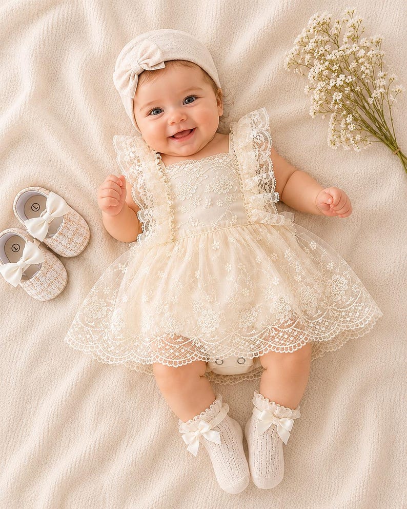 May include: A smiling baby wearing a cream-colored lace dress, matching headband, and socks with bows. The baby is also wearing a pair of cream-colored shoes with bows. A bouquet of white flowers is next to the baby.