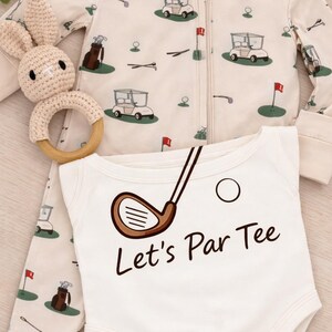May include: A baby outfit set. The cream-coloured onesie has the text "Let's Par Tee" and a golf club graphic. The set includes a matching zip-up sleeper with golf course illustrations and a crochet bunny rattle. baby gift for new golf loving parents. baby gift for golfer. daddy baby announcement golf. daddy golfing baby. golf and baby