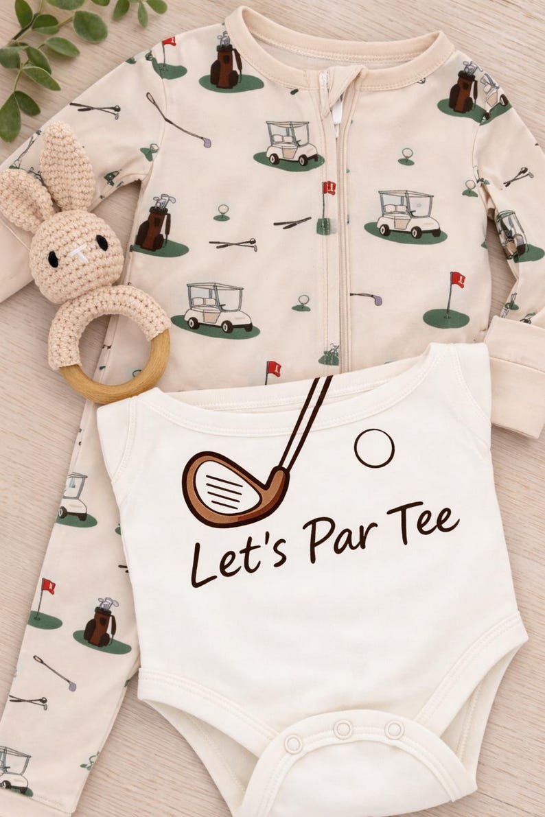 May include: A baby outfit set. The cream-coloured onesie has the text "Let's Par Tee" and a golf club graphic. The set includes a matching zip-up sleeper with golf course illustrations and a crochet bunny rattle. baby gift for new golf loving parents. baby gift for golfer. daddy baby announcement golf. daddy golfing baby. golf and baby
