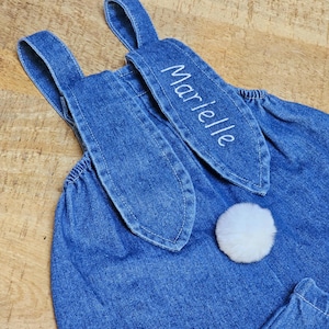 A blue denim overall with bunny ear detailing. The ears are embroidered with the name "Marielle" in white. A fluffy white pom-pom is attached to the back, resembling a bunny tail. The overall is designed for a child. Cute bunny Easter overall