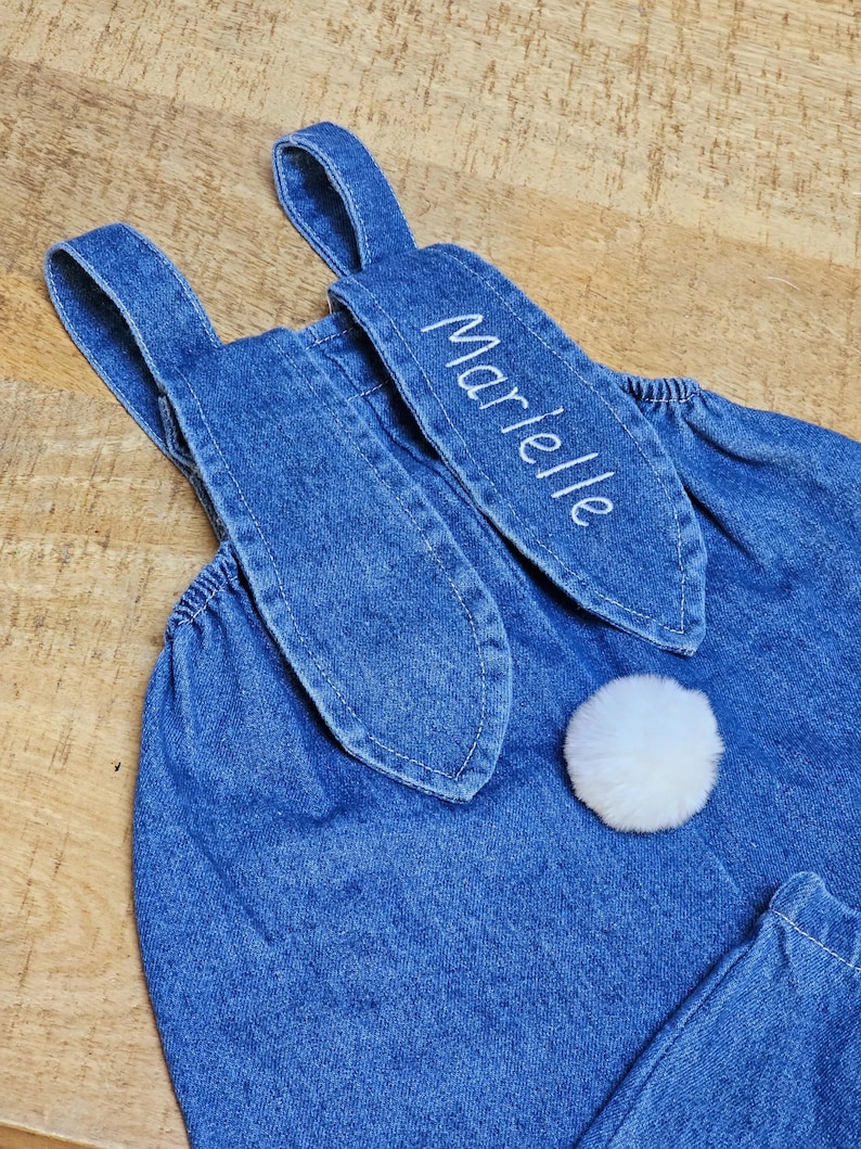 A blue denim overall with bunny ear detailing. The ears are embroidered with the name "Marielle" in white. A fluffy white pom-pom is attached to the back, resembling a bunny tail. The overall is designed for a child. Cute bunny Easter overall