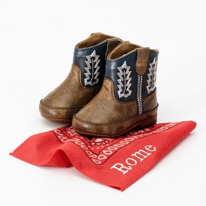 Pode incluir: Um par de botinhas de cowboy castanhas e azul-marinho para beb&eacute; com costuras brancas, sobre um len&ccedil;o vermelho com o nome "Rome" impresso em branco. As botinhas t&ecirc;m um design western cl&aacute;ssico.