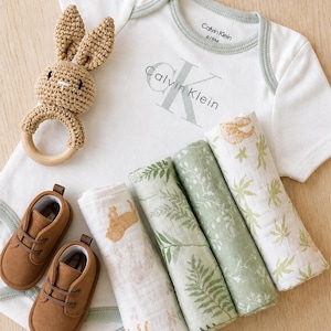 May include: A selection of baby essentials: a white Calvin Klein onesie, a crochet bunny rattle, brown baby shoes, and rolled muslin cloths with botanical and animal patterns. The onesie is size 6/9M. The items are arranged on a light-colored surface. branded baby boy gift set. baby boy hamper set. baby boy Calvin Klein set. baby boy Tommy Hilfiger se
