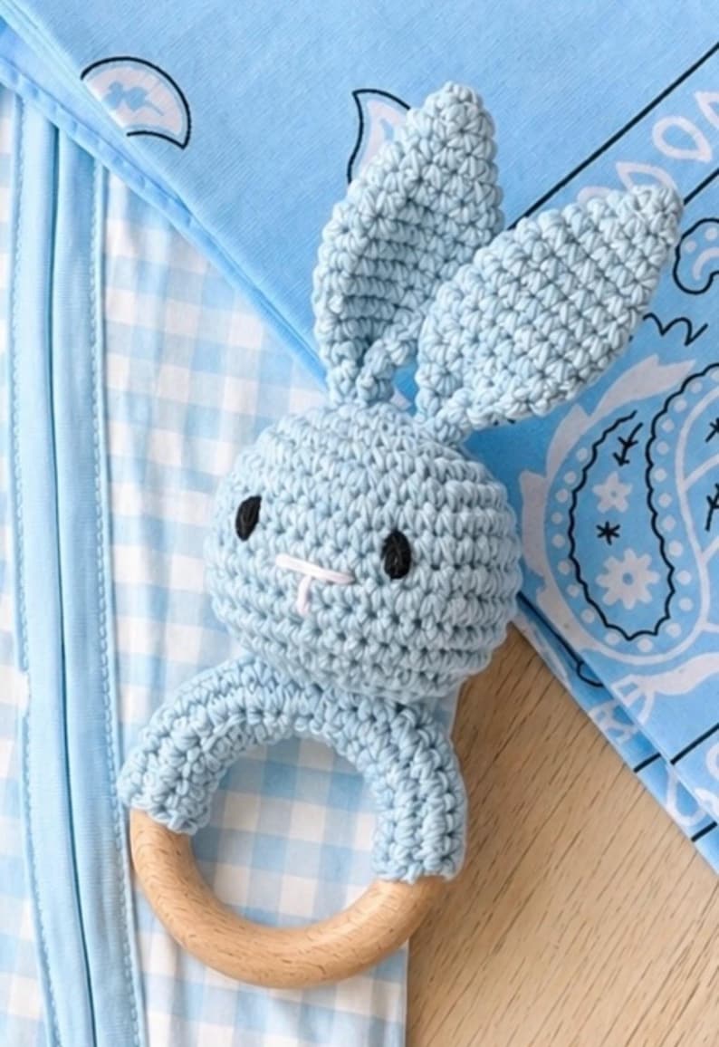 May include: A light blue crochet bunny rattle with a wooden ring base. The bunny has black eyes and a white stitched mouth. The rattle is on a blue and white checkered background with a blue paisley pattern.