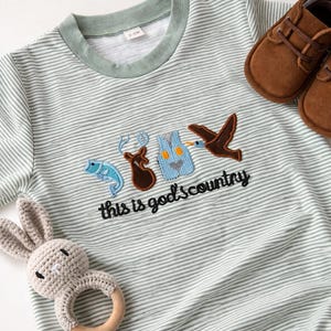 May include: A light green and white striped baby onesie with the text "this is god's country" and embroidered patches of a fish, deer, vest, and bird. Includes brown baby shoes and a crochet bunny rattle.