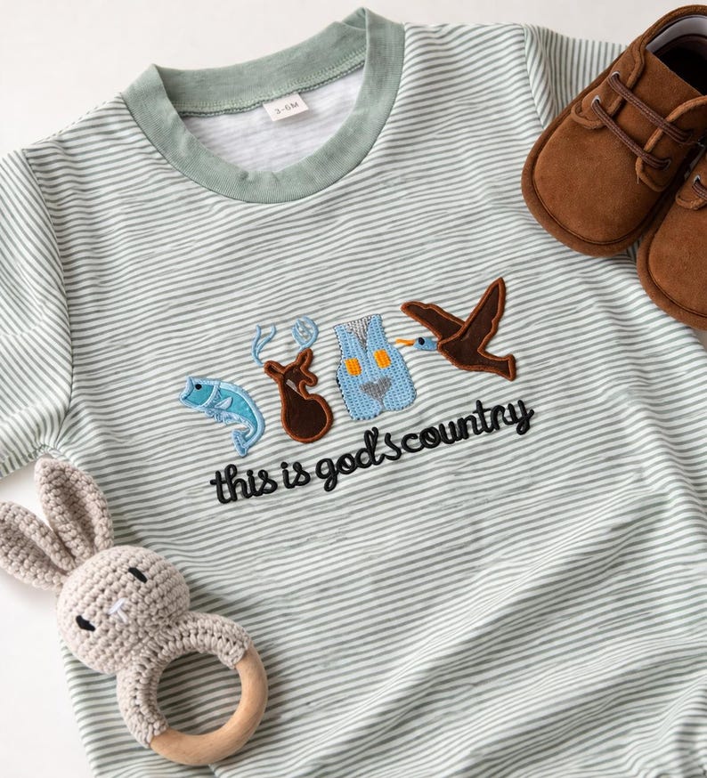 May include: A light green and white striped baby onesie with the text "this is god's country" and embroidered patches of a fish, deer, vest, and bird. Includes brown baby shoes and a crochet bunny rattle.