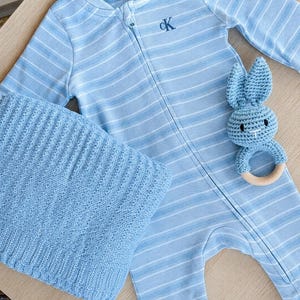 May include: A baby blue striped onesie with a zipper, a matching knitted blanket, and a blue crochet bunny rattle with a wooden ring. The onesie has the letters "CK" embroidered on the chest.