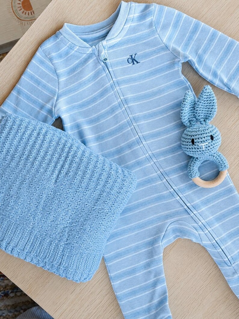 May include: A baby blue striped onesie with a zipper, a matching knitted blanket, and a blue crochet bunny rattle with a wooden ring. The onesie has the letters "CK" embroidered on the chest.