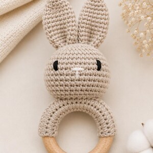 May include: A beige crochet bunny rattle with a wooden ring base. The bunny has black eyes and a white stitched nose. The ears are upright. The rattle is designed for babies and young children.