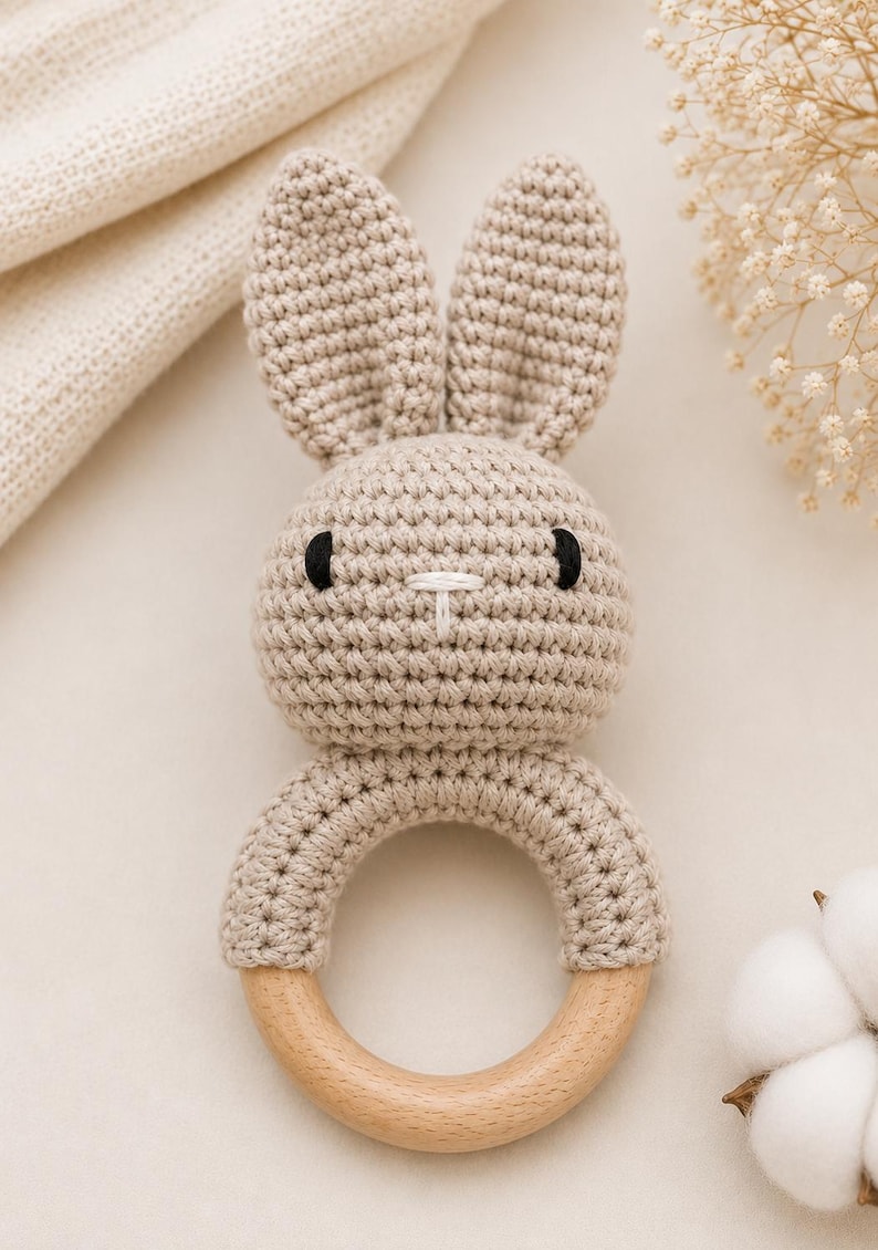 May include: A beige crochet bunny rattle with a wooden ring base. The bunny has black eyes and a white stitched nose. The ears are upright. The rattle is designed for babies and young children.