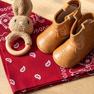 May include: A pair of tan baby cowboy boots with white stitching, next to a crocheted bunny rattle with a wooden ring. The items rest on a red bandana with a white paisley pattern. texas baby gifts. Michigan baby gifts. Montana baby gifts. southern baby gifts.