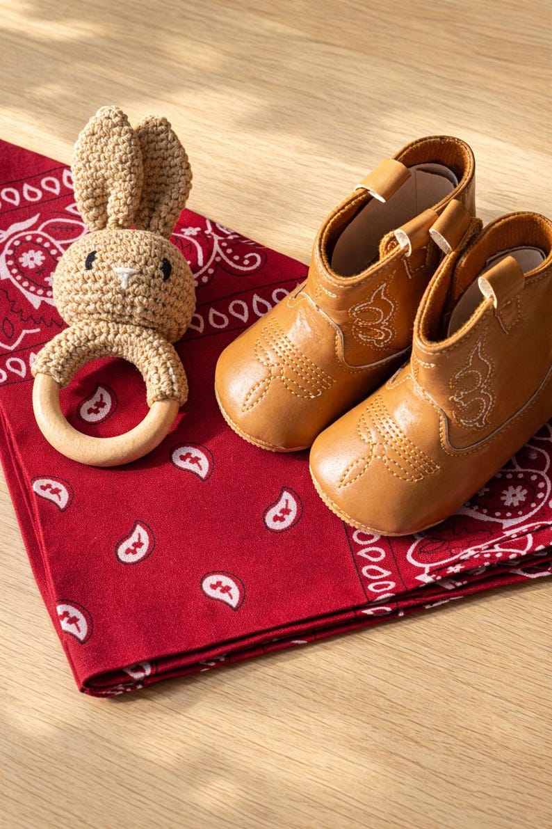 May include: A pair of tan baby cowboy boots with white stitching, next to a crocheted bunny rattle with a wooden ring. The items rest on a red bandana with a white paisley pattern. texas baby gifts. Michigan baby gifts. Montana baby gifts. southern baby gifts.