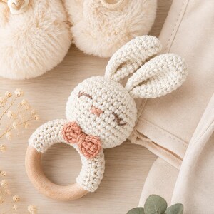 May include: A handmade crochet bunny rattle with a wooden ring. The bunny has cream-colored yarn with a pink bow and embroidered facial features. The rattle is part of a baby set, with matching cream-colored baby booties.