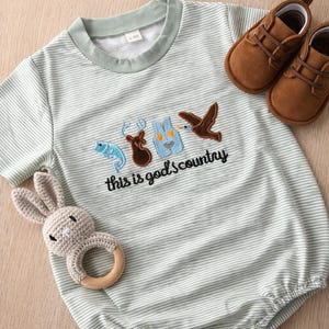May include: A light green and white striped baby romper with embroidered images of a fish, deer, vest, and bird, with the text "this is god's country". Includes a brown pair of baby shoes and a gray crochet bunny rattle.