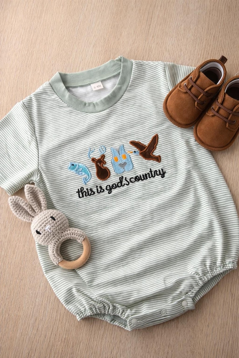 May include: A light green and white striped baby romper with embroidered images of a fish, deer, vest, and bird, with the text "this is god's country". Includes a brown pair of baby shoes and a gray crochet bunny rattle.
