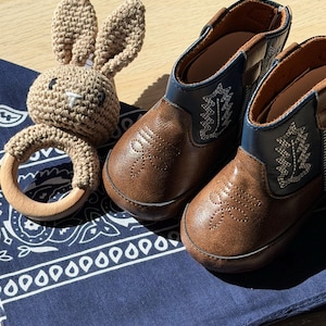 May include: A pair of brown and blue faux leather boots with white stitched leaf patterns and a crochet bunny rattle featuring a light brown yarn head and ears attached to a smooth, round, light-colored wooden ring, all resting on a navy blue bandana.