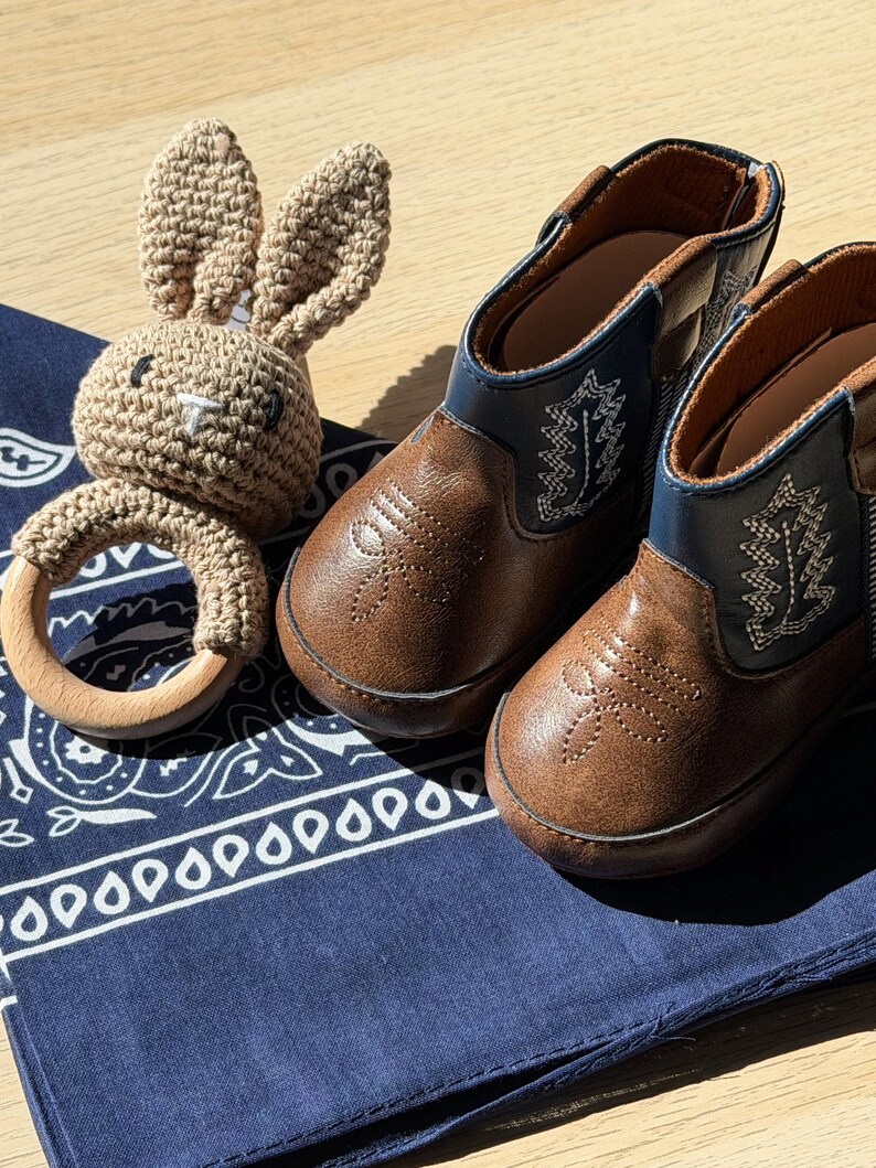 May include: A pair of brown and blue faux leather boots with white stitched leaf patterns and a crochet bunny rattle featuring a light brown yarn head and ears attached to a smooth, round, light-colored wooden ring, all resting on a navy blue bandana.