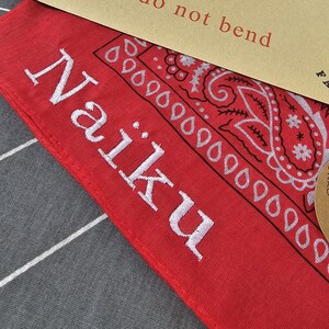 May include: Red bandana with white embroidered text "Naiku" and a paisley design. The bandana is folded, with a portion of a brown paper tag visible in the upper right corner, which reads "do not bend".