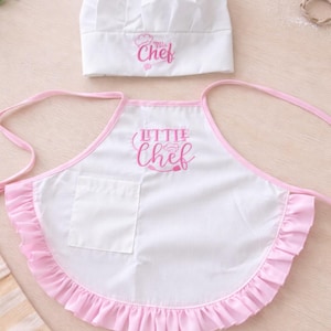 A baby child's chef's hat and apron set. The apron has a pink frilled trim and pocket. The words "Little Chef" are printed in pink on the apron and hat. The set is designed for children. baby gift ideas for a chef