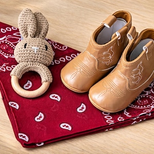 Tan baby cowboy boots with white stitching, a crochet bunny teether, and a red bandana with a white paisley pattern. Baby boy cowboy boots gift set. baby girl cowgirl boots gift set. cowboy baby gift set ideas. Western themed baby shower gift. Texas baby gift ideas. country baby gift ideas. baby boy gift ideas. baby girl gift ideas affordable. baby boy gift ideas adorable. newborn gift ideas. newborn gift set. baby cowboy boots. red paisley baby cowboy. baby cowboy hat. texas baby gift.