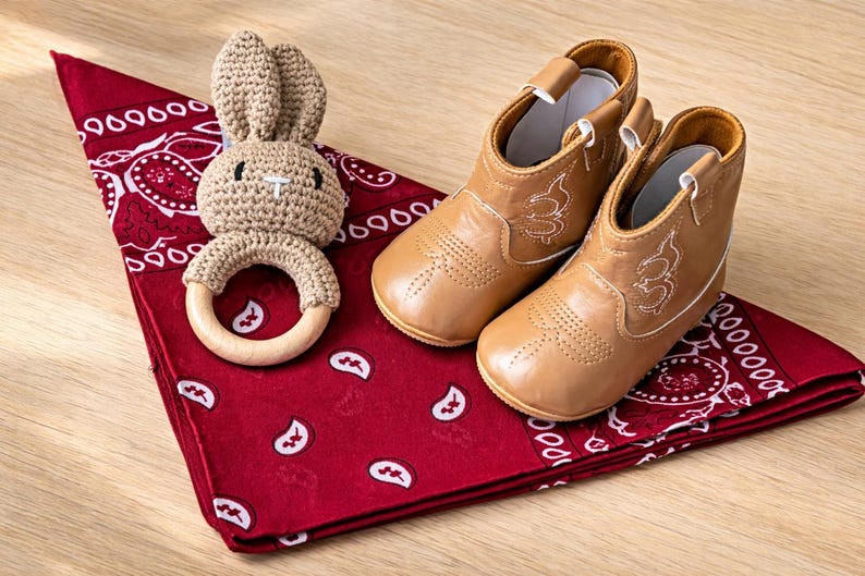 Tan baby cowboy boots with white stitching, a crochet bunny teether, and a red bandana with a white paisley pattern. Baby boy cowboy boots gift set. baby girl cowgirl boots gift set. cowboy baby gift set ideas. Western themed baby shower gift. Texas baby gift ideas. country baby gift ideas. baby boy gift ideas. baby girl gift ideas affordable. baby boy gift ideas adorable. newborn gift ideas. newborn gift set. baby cowboy boots. red paisley baby cowboy. baby cowboy hat. texas baby gift.