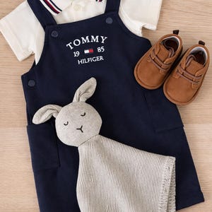 A navy blue baby outfit set with a white polo shirt, navy blue overalls with the "TOMMY HILFIGER" logo, brown baby shoes, and a gray bunny comforter. The set is designed for infants. Baby boy luxury branded gift set in ready gifted box. Baby gift set ideas for baby boy baby shower