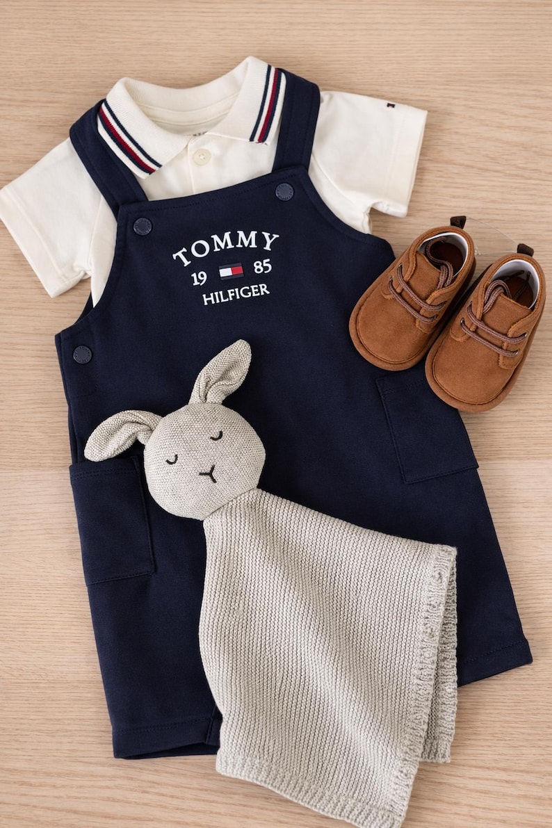 A navy blue baby outfit set with a white polo shirt, navy blue overalls with the "TOMMY HILFIGER" logo, brown baby shoes, and a gray bunny comforter. The set is designed for infants. Baby boy luxury branded gift set in ready gifted box. Baby gift set ideas for baby boy baby shower
